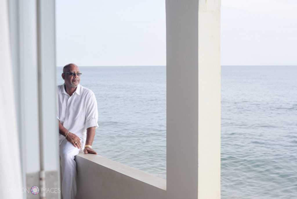 The groom sits on the edge of the porch over looking the ocean he is wearing all white clothes and sunglasses.