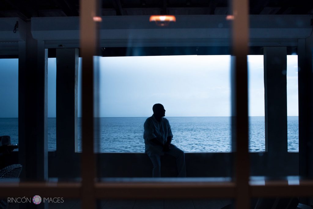Sihouette photograph of the groom sitting on the edge of the porch railing with the blue ocean in the background.